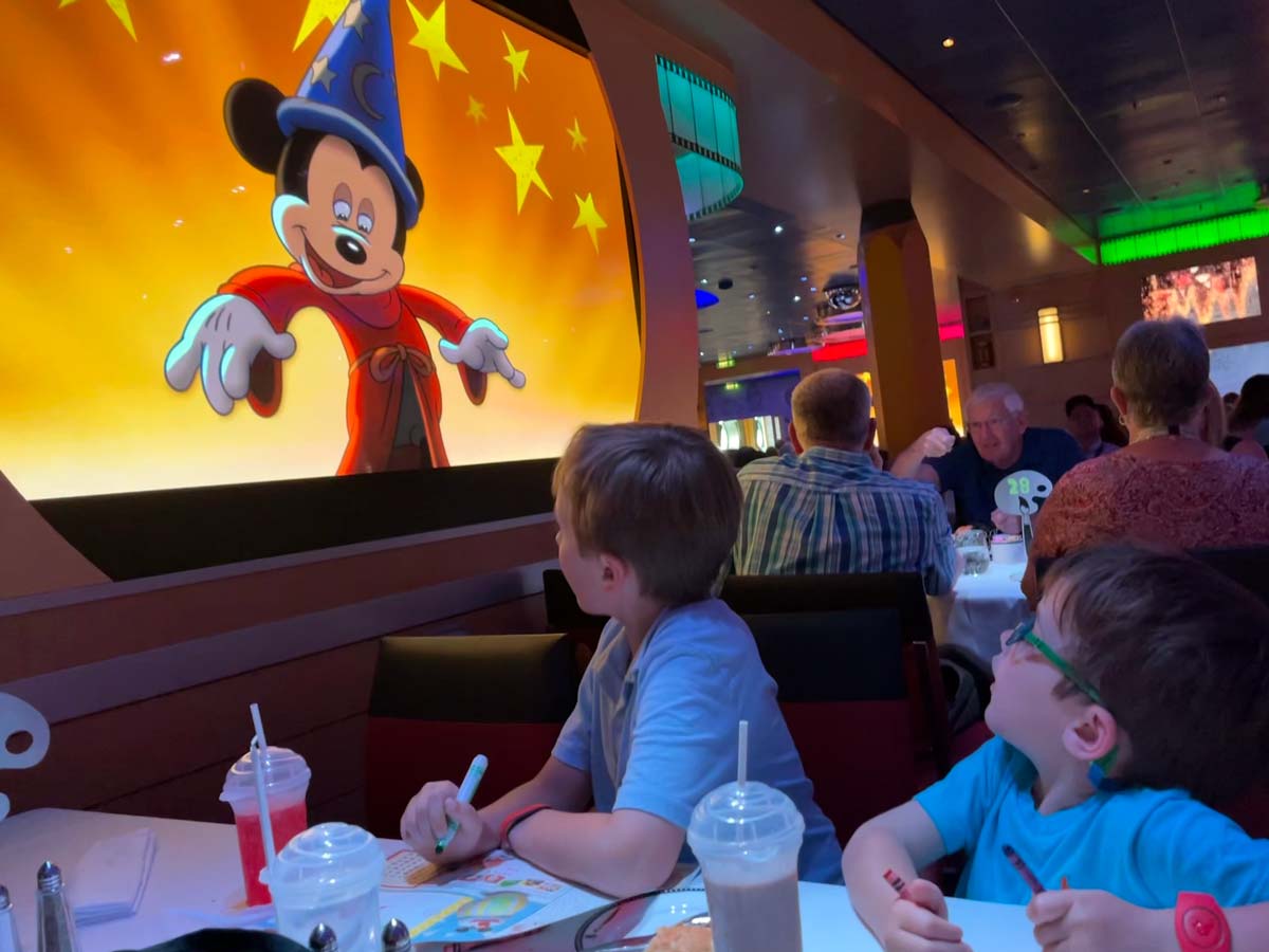 Two young boys look up at a screen, where Mickey Mouse is dancing, while eating dinner on a Disney Cruise Line.