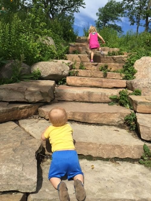 A young girl walks, and a baby boy crawls, up stone stairs in Madison's Picnic Point, one of the best affordable summer vacations in the United States with kids.