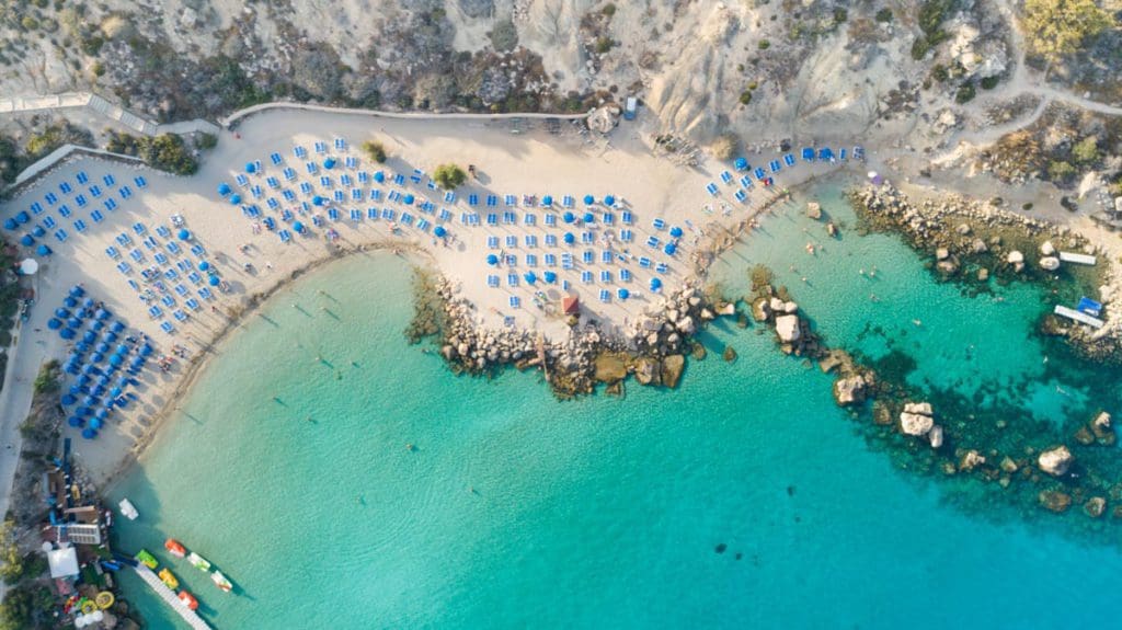 An aerial view of several blue umbrellas lined up along Konos Beach in Cyprus, one of the best beach destinations in Europe for families.
