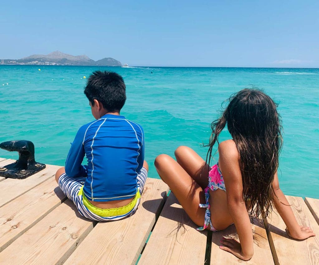 Two kids sit on a dock looking out onto a clear blue ocean in Mallorca.