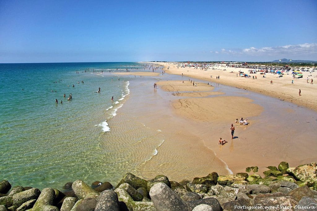 A long stretch of beach in Tavira, dotted with people on a sunny day.