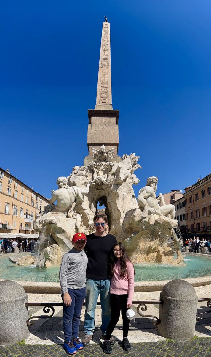 A dad and his two kids stand in front of a Roman fountain during the day.