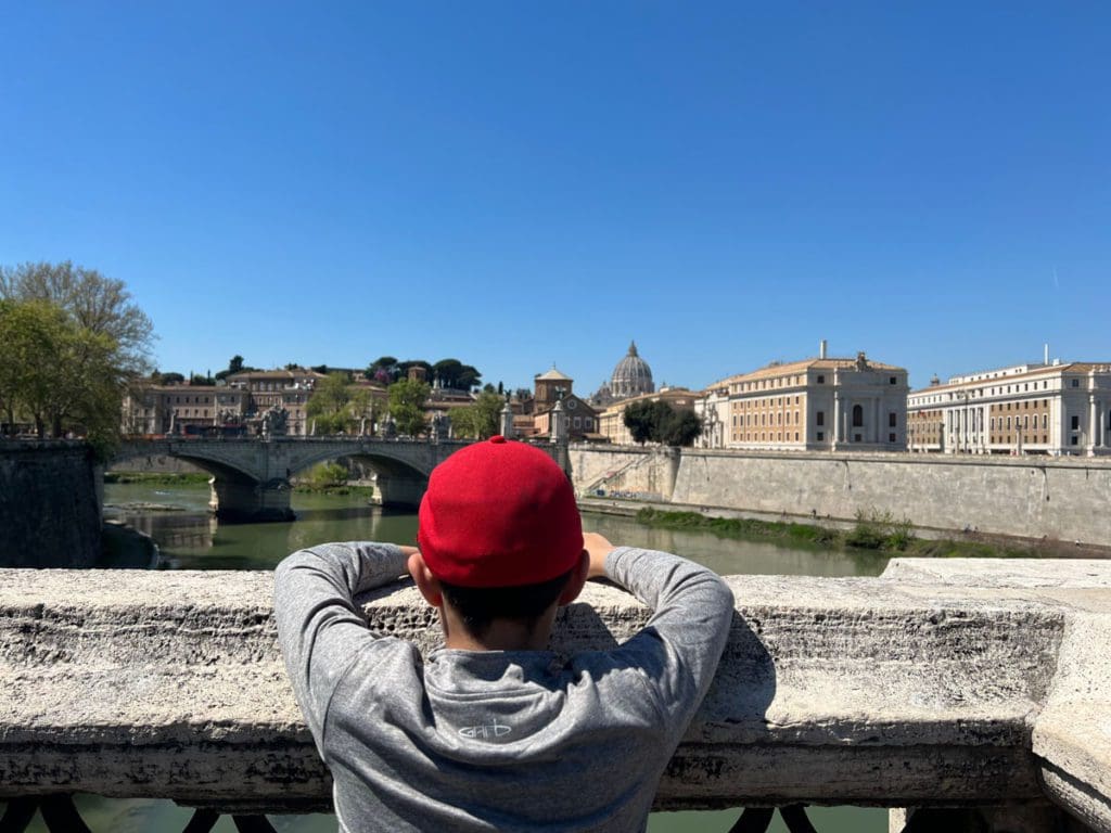 A young boy looks over a bridge at the river in Rome, as well as the surrounding sights.