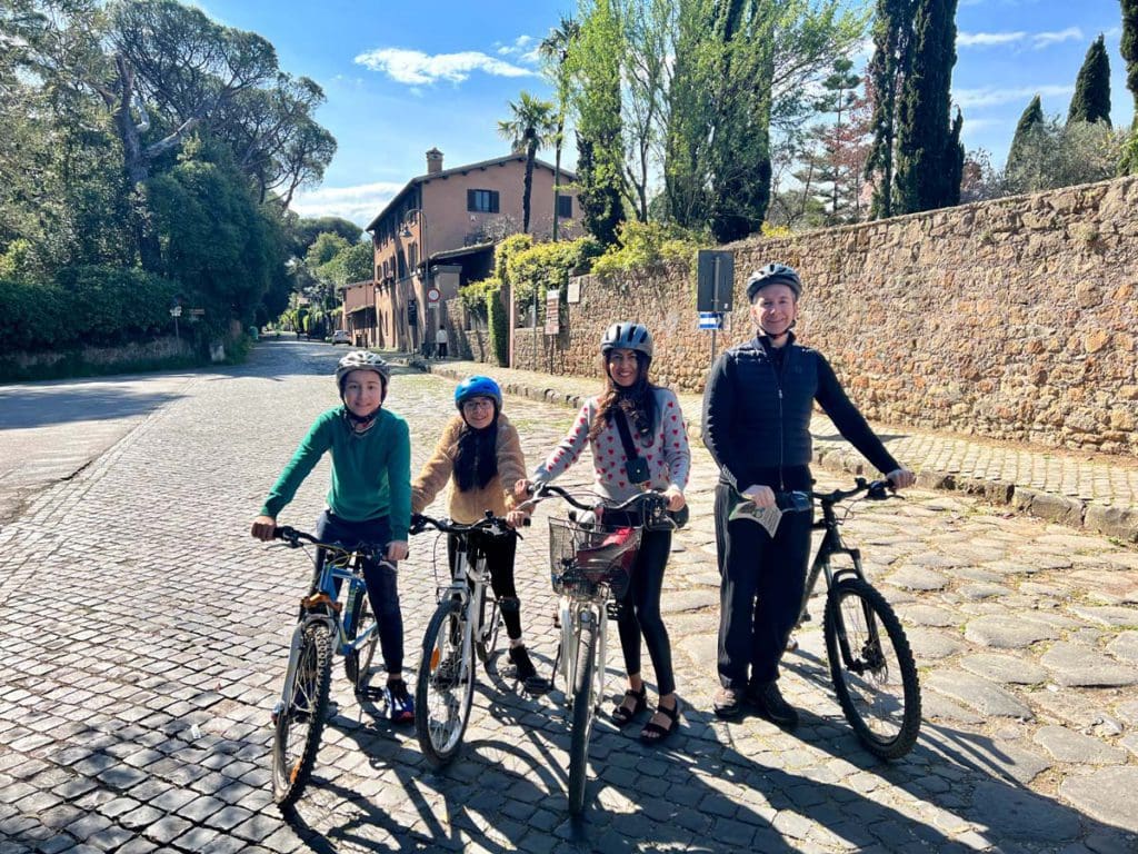 A family of four, atop their own bikes, poses along a stretch of road on the Appian Way, learning about things to do in Rome is an important part of knowing all about Italy with kids.