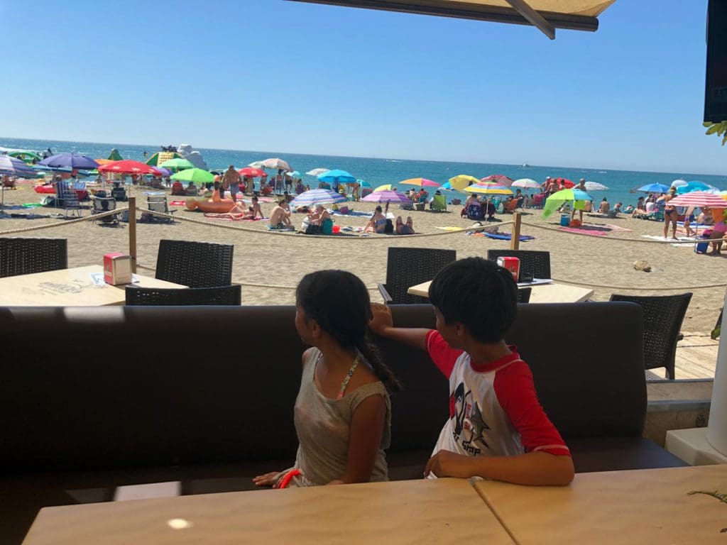 Two kids turn around in their chairs to look at the crowded beach of Costa del Sol, one of the best beach destinations in Europe for families.