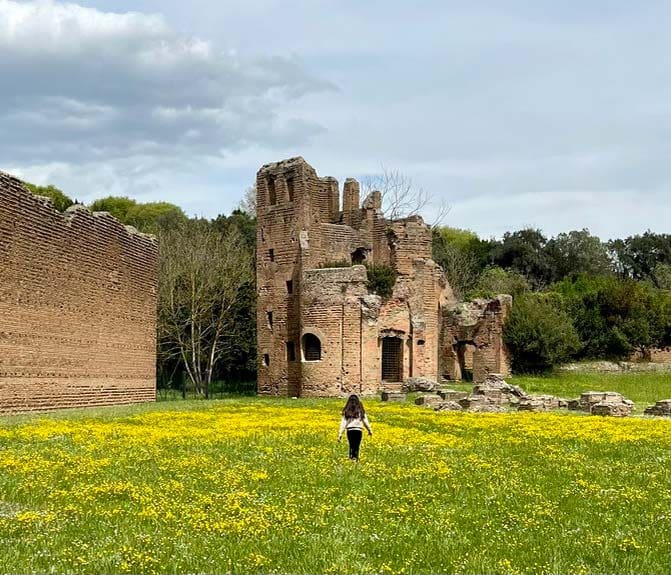 A young girl runs through a yellow filed, with Roman ruins in the distance, while exploring the Appian Way, a must stop on our Rome itinerary with kids.