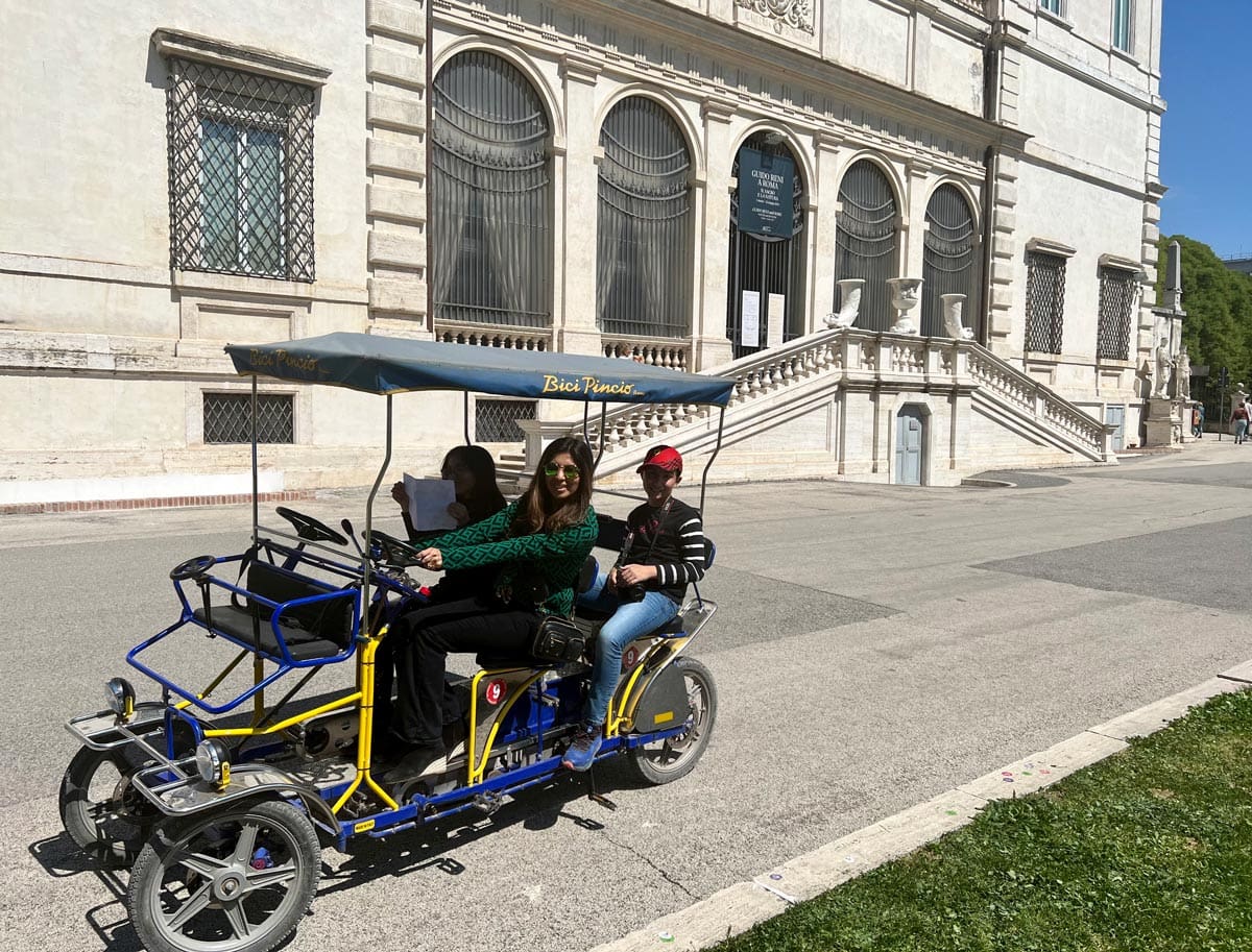 A family of four rides a surrey bike around Villa Borghese, with the museum behind them.