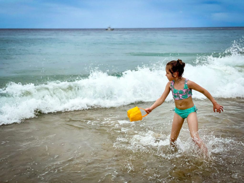 A young girl wearing a swim suit, holding a yellow bucket, plays in the waves on the sand at Crash Boat Beach in Puerto Rico, one of the best affordable Caribbean islands for families.