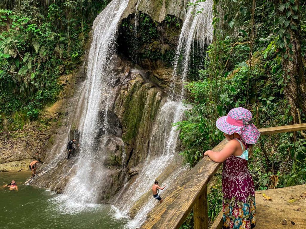 A young girl stands on a platform observing the waterfall and pool below at Gozalandia Waterfall, where others are swimming in the water.