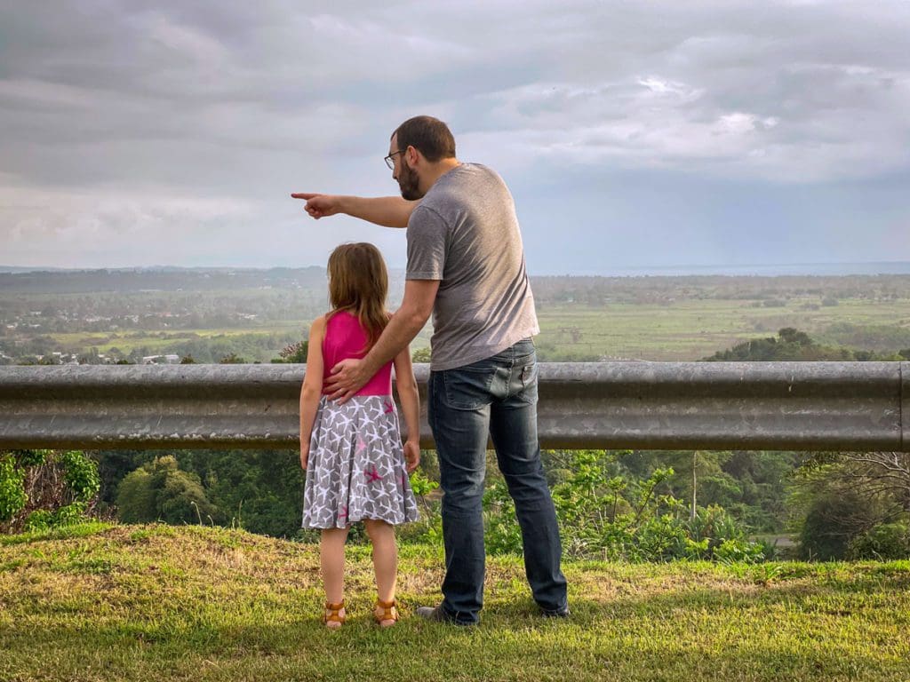 A dad stands with his young daughter, while pointing for her to see something in the distance with a view of Puerto Rico beyond them.
