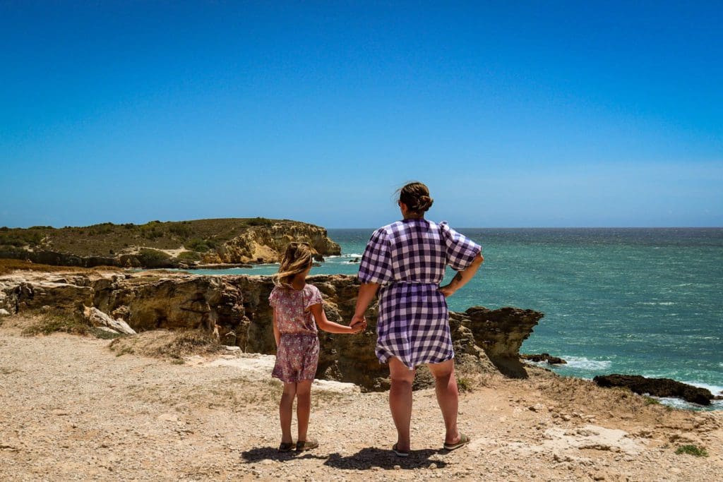 A mom and her young daughter stand hand-in-hand while enjoying a view of the ocean from the cliffs of Cabo Rojo National Wildlife Refuge In Puerto Rico, one of the best affordable Caribbean islands for families 