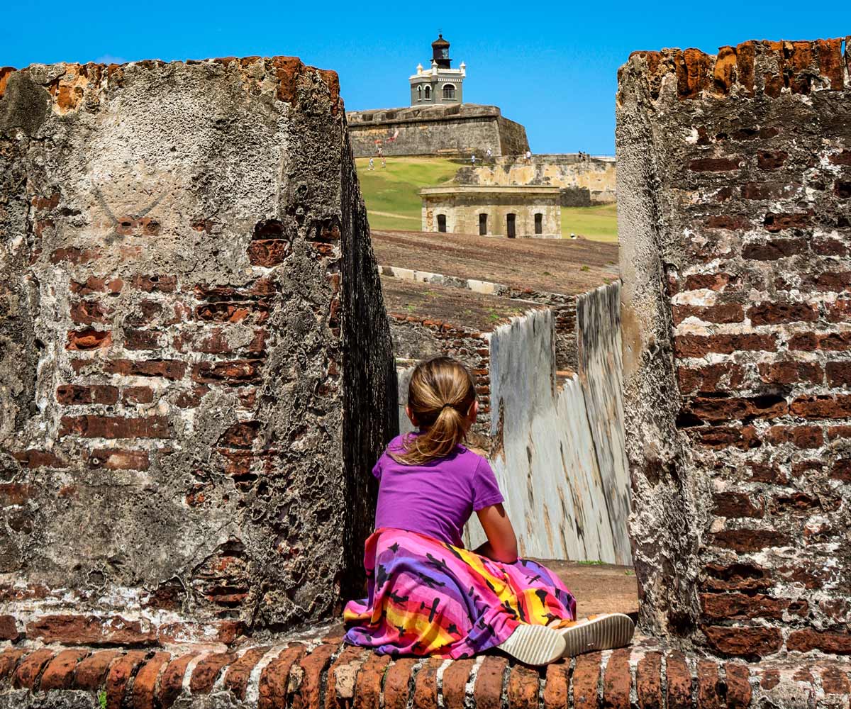 A young girl sits along a brick wall looking at San Felipe del Morro Castle in San Juan.