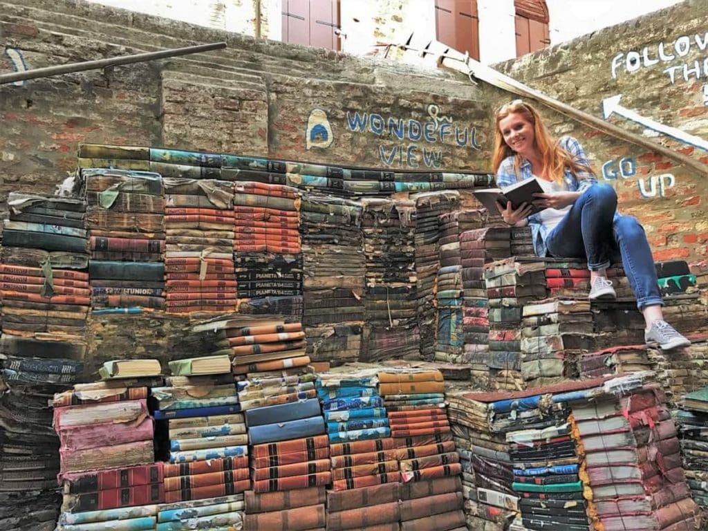 A woman leans against an outdoor display of books at the iconic Acqua Alta Bookshop.
