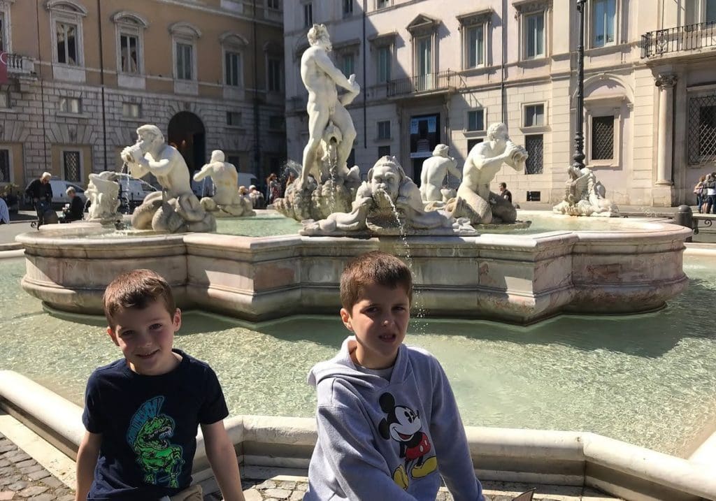 Two pose sit at the edge of a fountain on one side of Piazza Navona. It's one of the best places to visit on an Italy itinerary with kids!