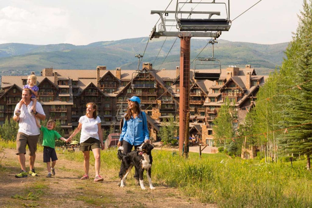 With a gondola in the background, a family with two children walks through the lush Colorado mountainside with a staff member and the resident dog of The Ritz-Carlton Beaver Creek.