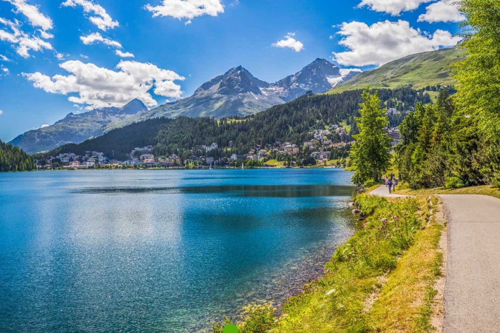 A road curves around a lake shoreline with mountains in the distance during the summer in St. Moritz.