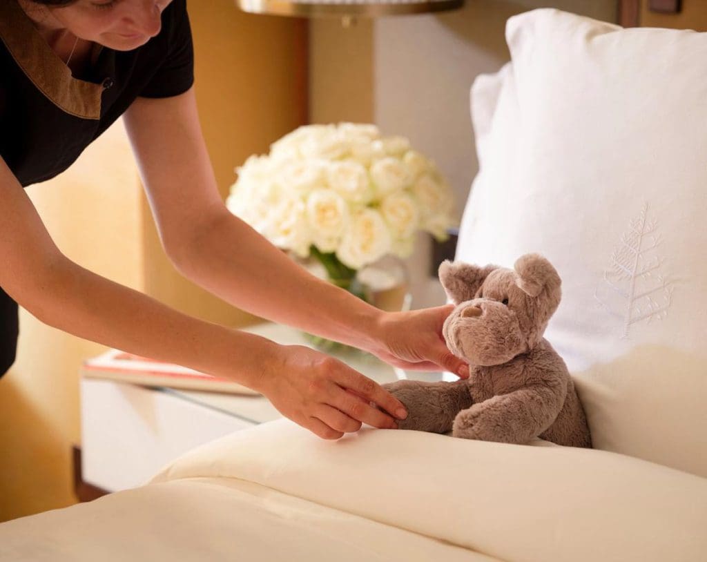 A hotel staff member tucks a teddy bear into a hotel bed at Four Seasons Hotel London at Park Lane, one of the best London Hotels for Families.