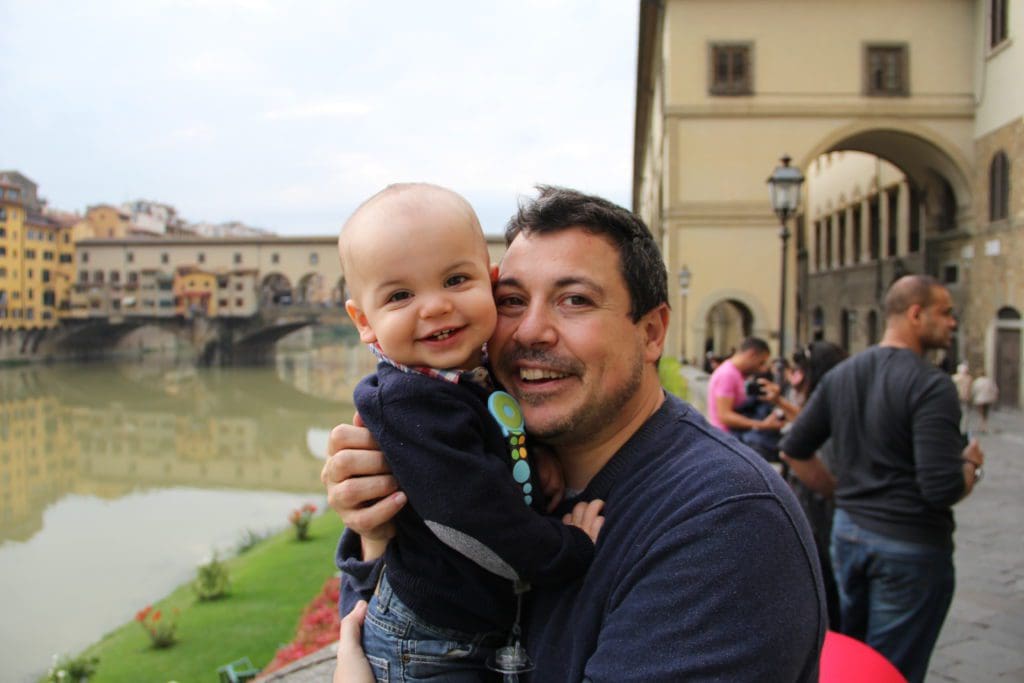 A dad holds a young baby with Ponte Vecchio in the distance, learning about things to do in Florence is an important part of knowing all about Italy with kids.