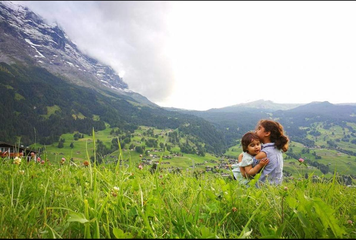 Two small kids embrace while sitting in a green pasture near Grindelwald, with a mountain view.