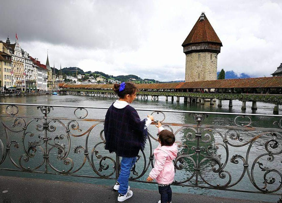 Two girls stand along a fence looking at the iconic lake and bridge in Lucerne, a must stop on any Switzerland itinerary with kids.