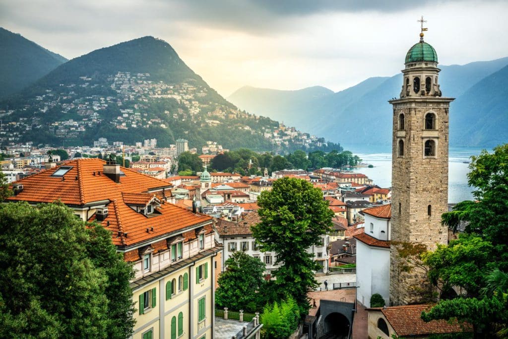 An aerial view of Lugano, feature a lake-side location, larger church tower, and orange-roofed buidlings.