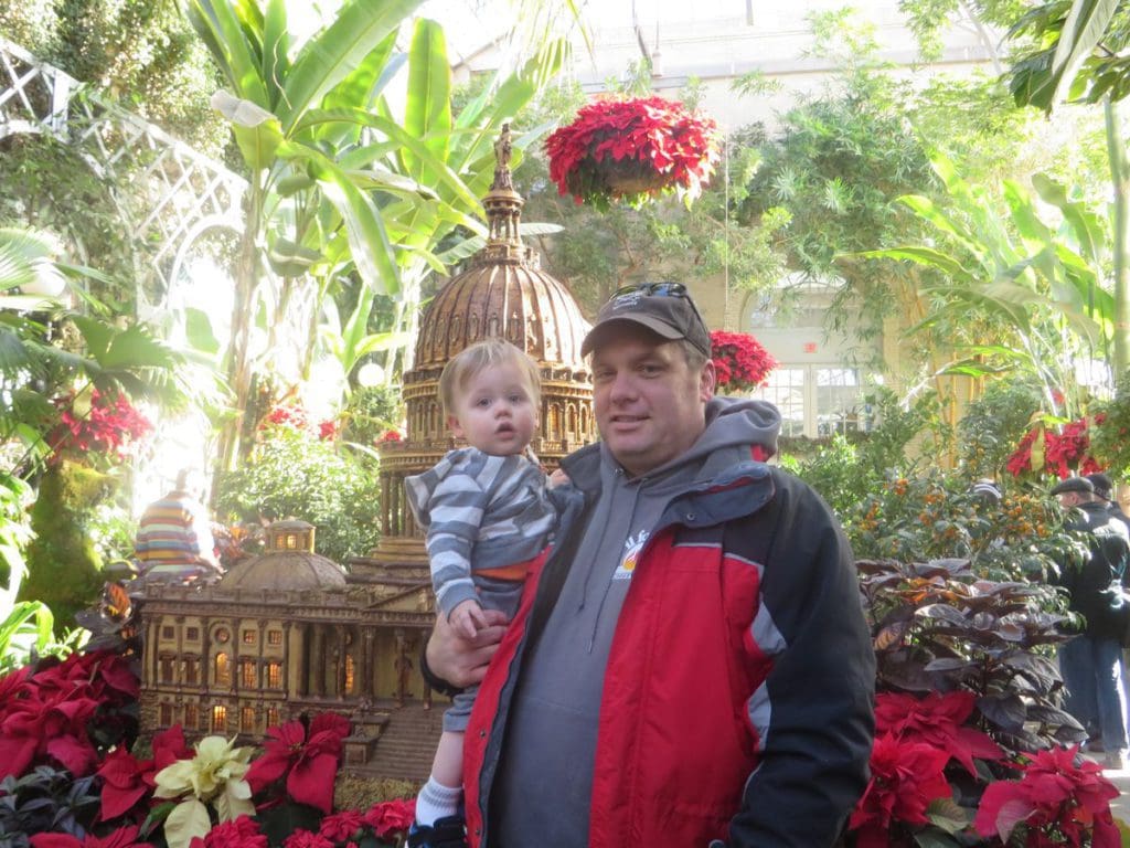 A dad holds his infant son with an array of foliage behind them at the US Botanical Garden in Washington DC.