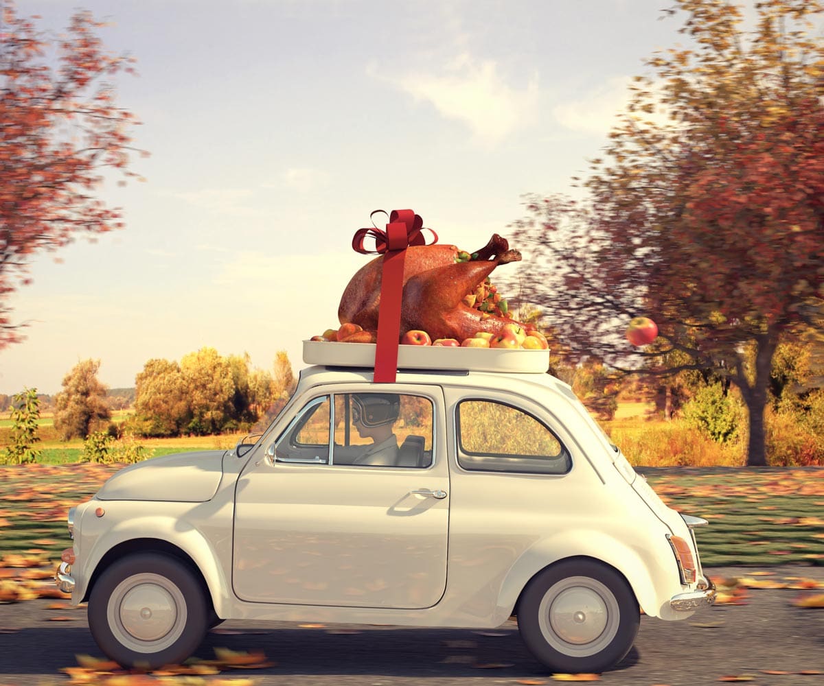 A small car drives through a fall landscape with a huge cooked turkey strapped to the roof.