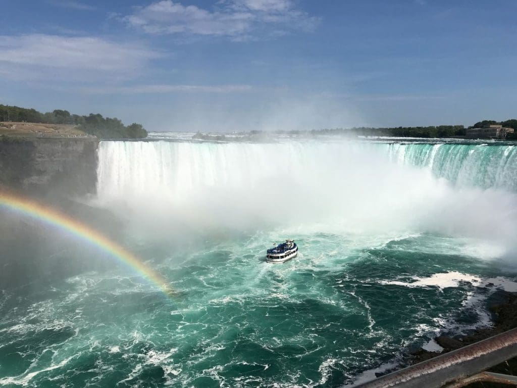 A rainbow arches toward Niagara Falls, while a a boat steers near the base of the falls, seen from the Canadian side.