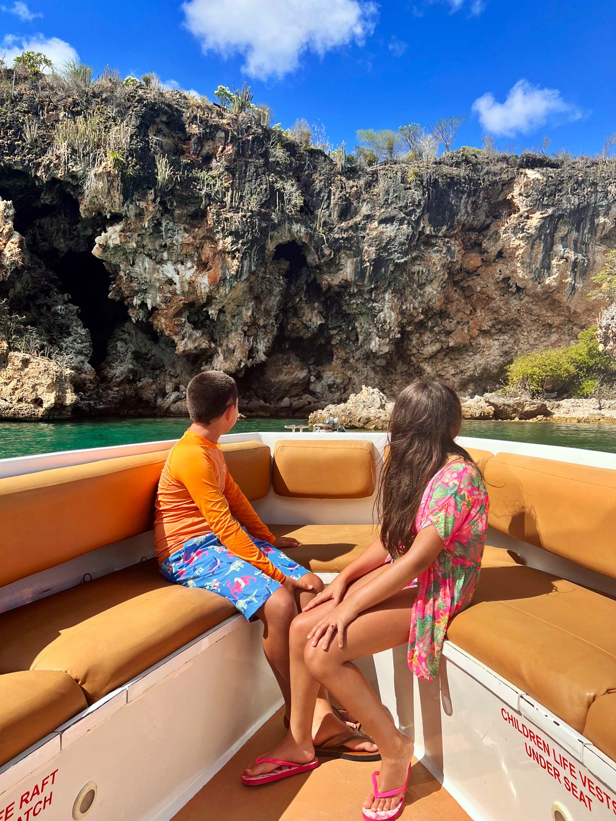 Two kids sit at the front of a boat looking at a rocky shoreline in Anguilla.