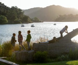 Several kids play near and on a large fallen log aside a lake at Alisal Ranch.