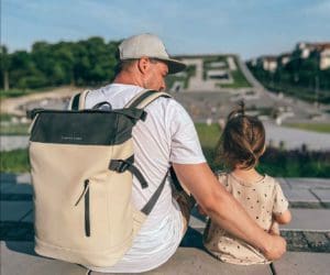 A dad, wearing a backpack, and his toddler daughter sit together taking in a city view.