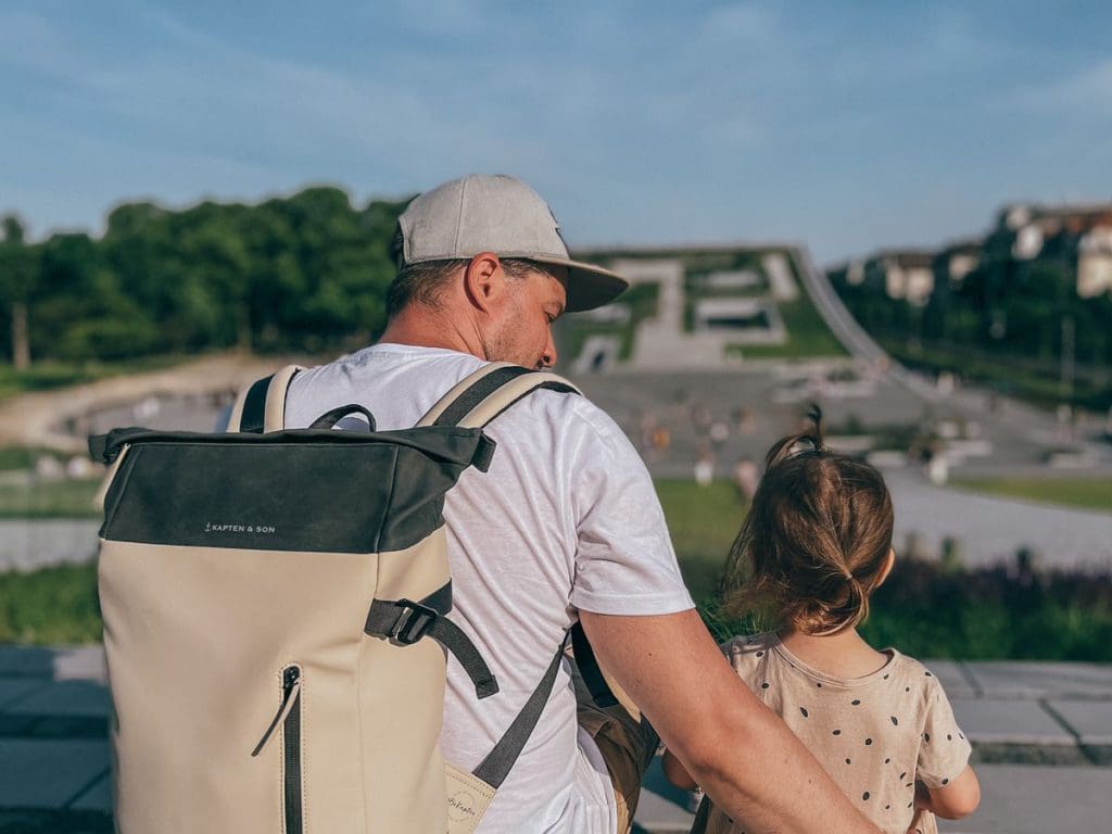 A dad, wearing a backpack, and his toddler daughter sit together taking in a city view - a backpack is a must-pack for a European vacation with toddlers.