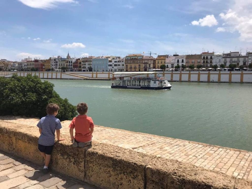 Two kids lean over a stone wall to see a boat floating down the river in Seville.