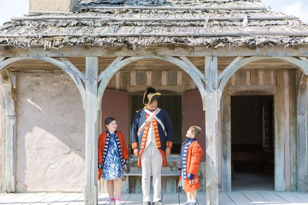 Two kids stand with a costumed reenact-or at Fort Michilimackinac.