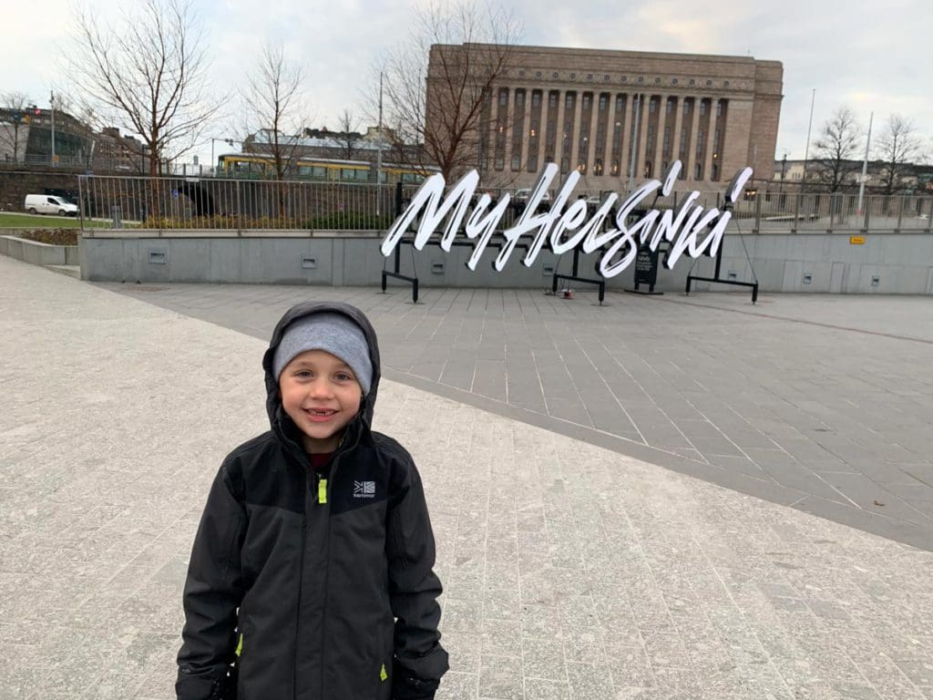 A young boy wearing a winter jacket and hat stands in front of a sign reading "My Helsinki", the first city on our Finland winter itinerary for families.