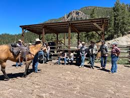 Several men wearing cowboy hats stand near a horse pen, while another leads a horse toward them at Rainbow Trout Ranch.