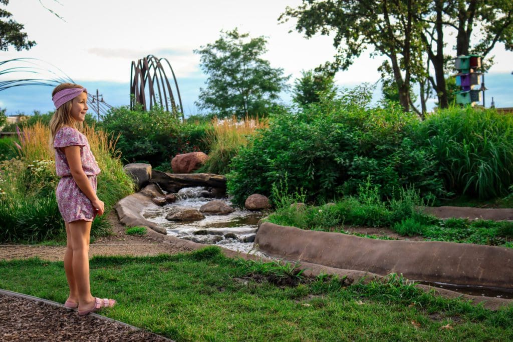 A young girl stands along a grassy area near a splash stream in River Prairie Park.