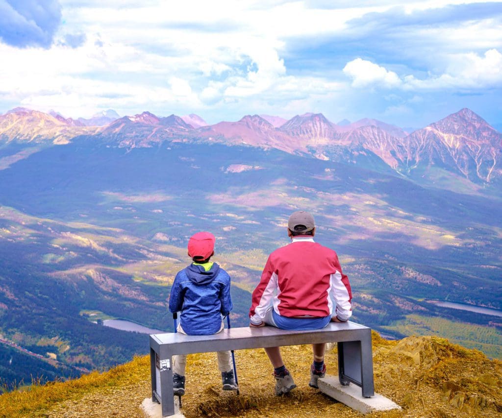 Father and son sit together atop a mountain on a beach with a sweeping view of the Canadian Rockies in front of them.