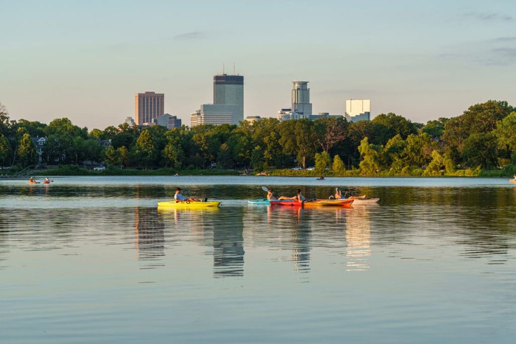 Several people paddle colorful kayaks on Lake of the Isles, with the Minneapolis skyline in the distance.