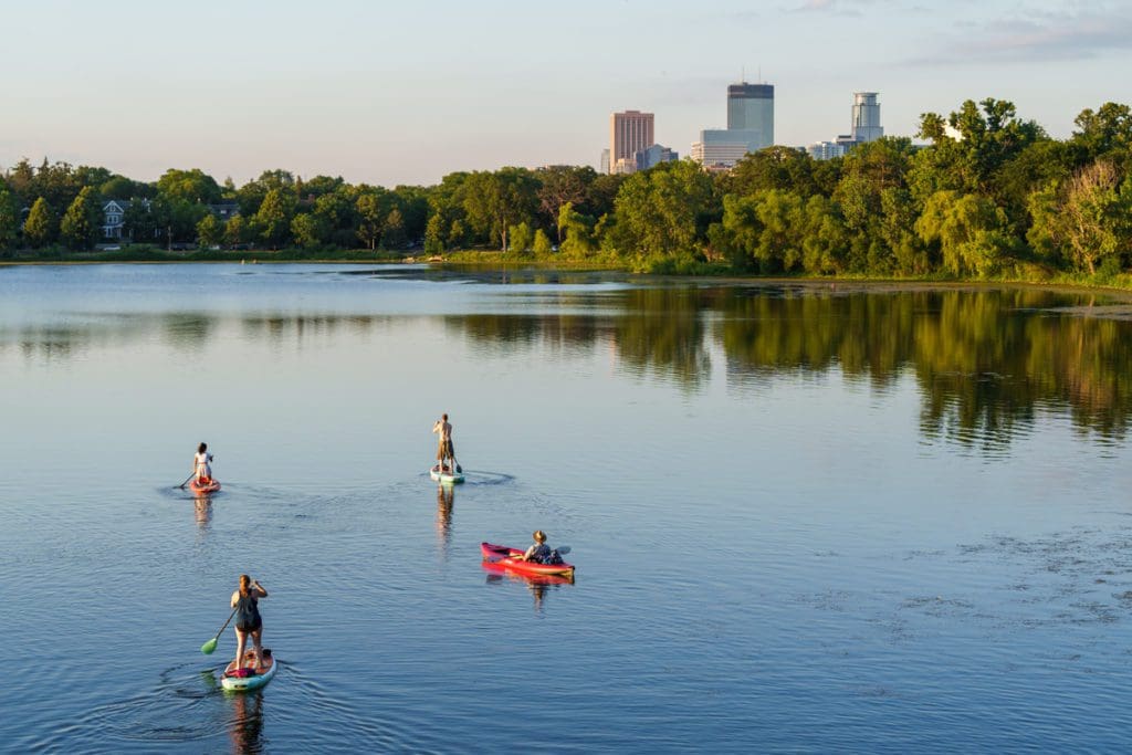 Four people paddle colorful kayaks around Lake of the Isles Park, with the Minneapolis skyline in the distance.