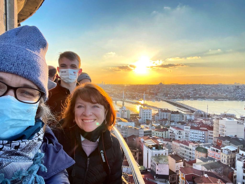 A family of three stands together and poses for a picture with a scenic view of Istanbul below them at sunset.