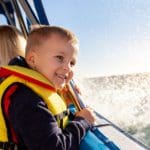 A little boy wearing a life jacket smiles broadly while waves splash onside of the sail boat.