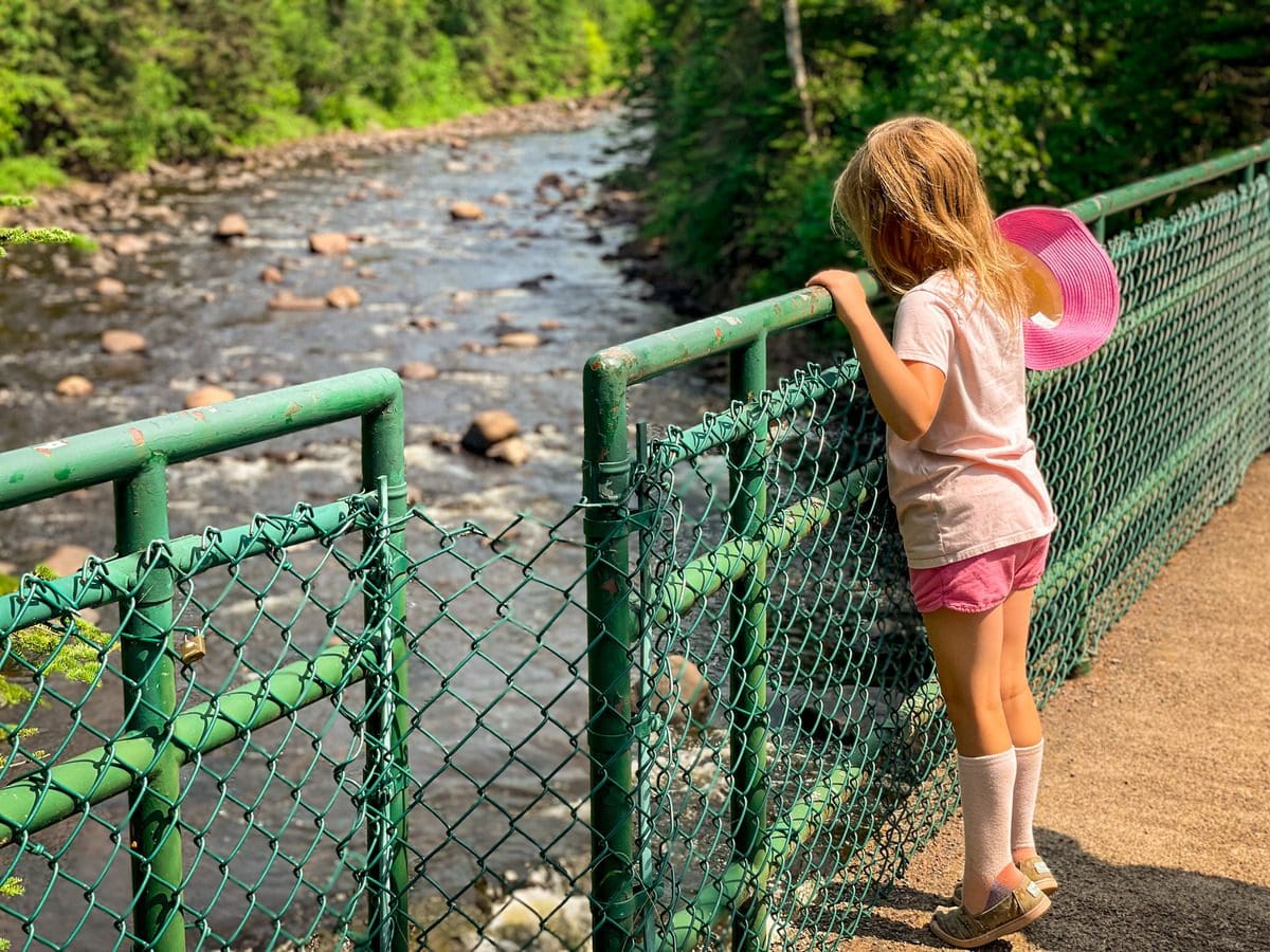 A young girl leans over a fence along a bridge, looking at the river below, in Judge C.R. Magney State Park, one of the best affordable summer vacations in the United States with kids is visiting a state park.