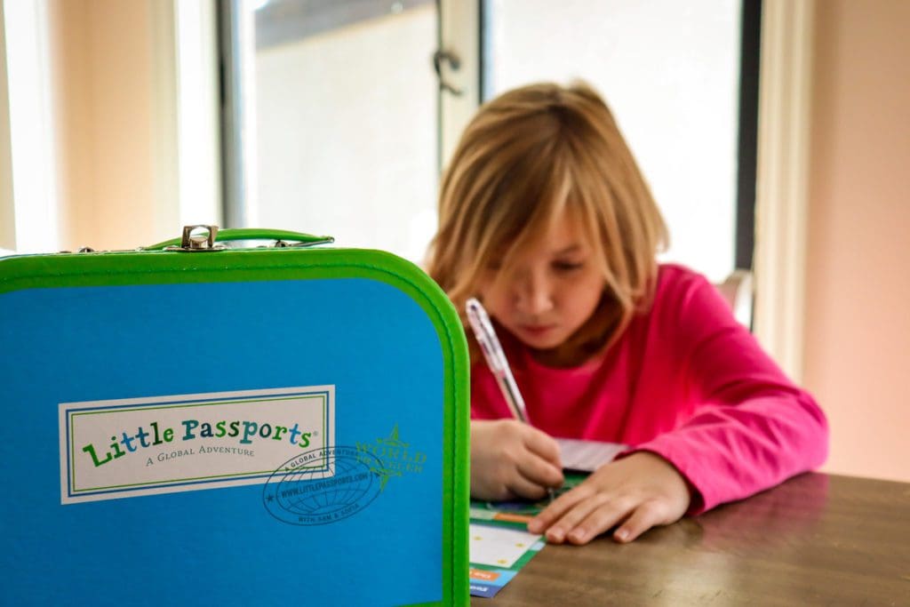 A Little Passports suitcase sits on a table, with a young girl behind it working on an activity.