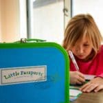 A Little Passports suitcase sits on a table, with a young girl behind it working on an activity.