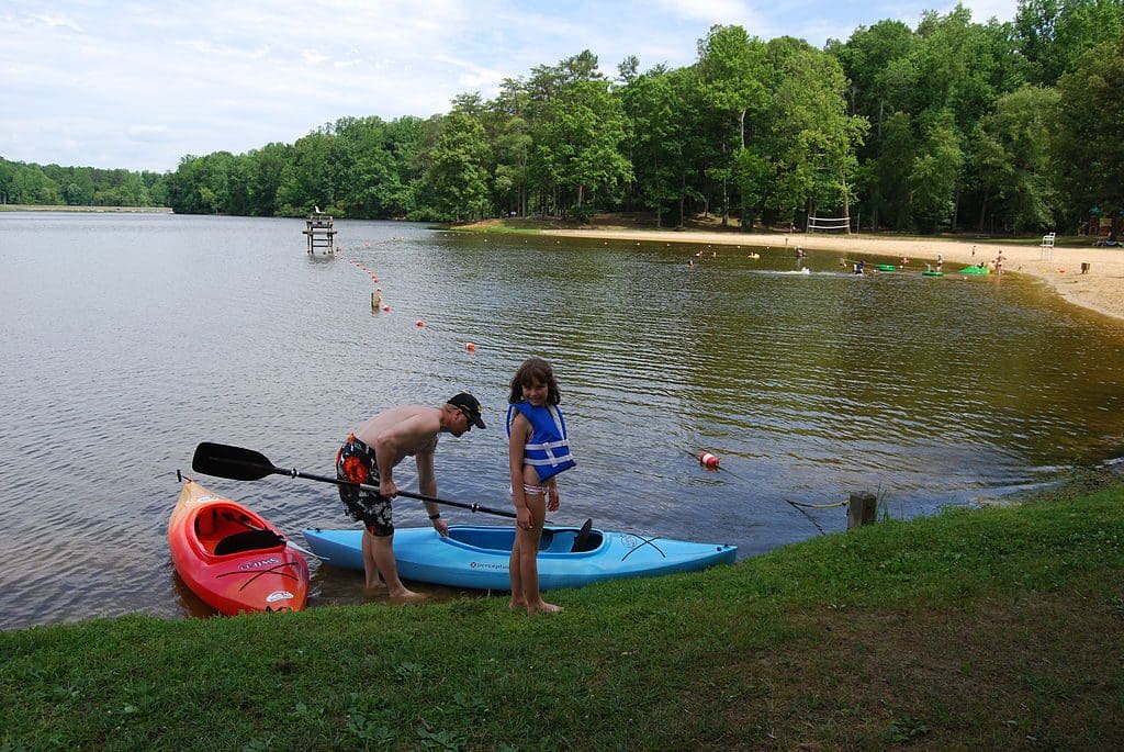 A child plays on the shores of Bear Creek Lake in Virginia, while dad brings in a kayak and a long stretch of beach rests behind them at one of the best lake getaways near Washington DC for families.