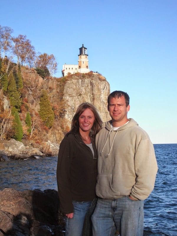 A man and woman stand together along the shore of Lake Superior, with Split Rock Lighthouse in the distance. 