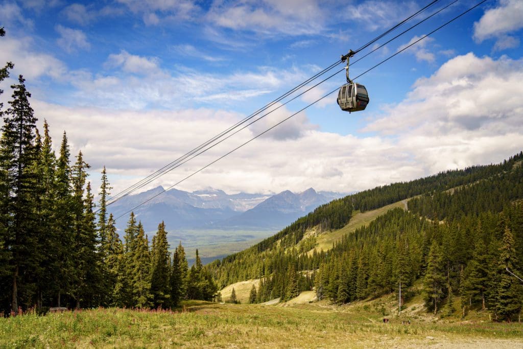 A gondola moves along the line with mountains surrounding it and Lake Louise in the distance.