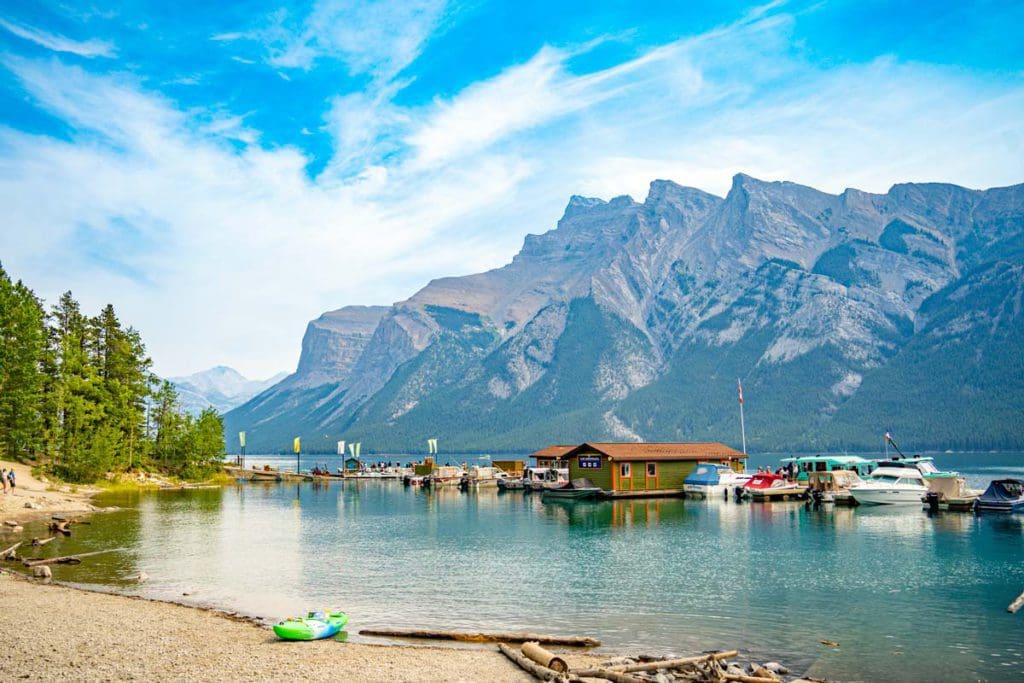 The shoreline of Lake Minnewanka, with boats gently floating off-shore and a view of the Canadian Rockies in the distance.
