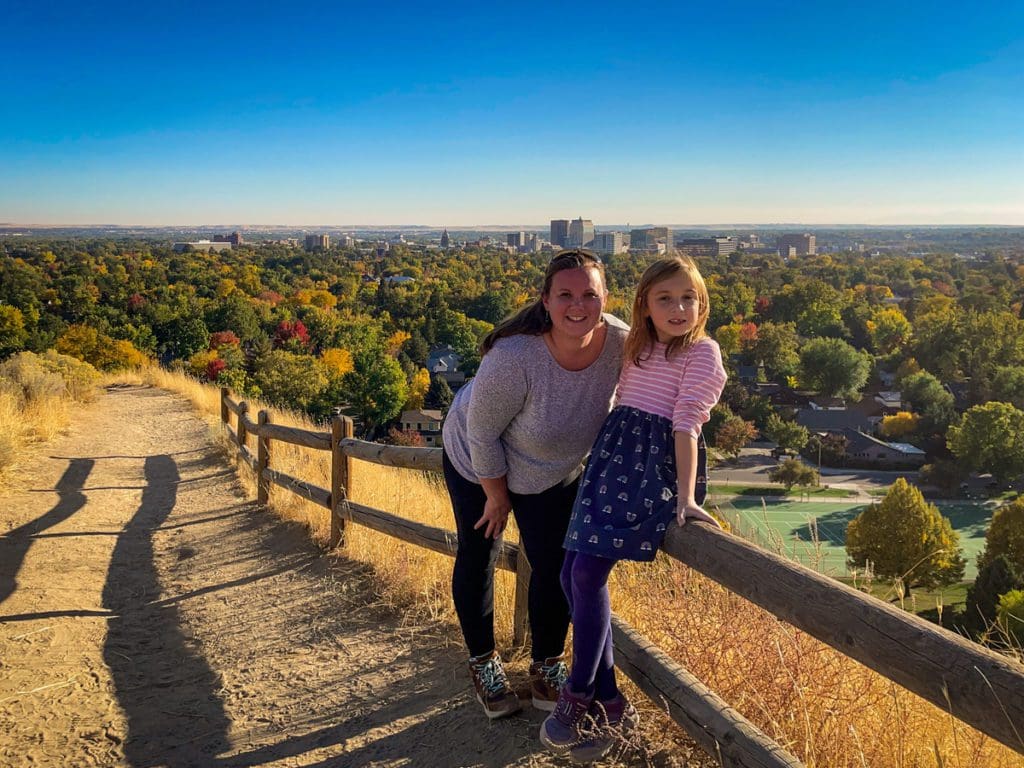 A mom and her daughter smile at the top of a Boise foothill, with an expansive view of downtown Boise in the distance, after hiking up a path at Camel's Back Park.